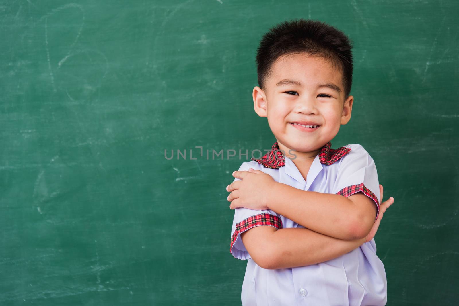 Back to School. Happy Asian funny cute little child boy from kindergarten in student uniform stand crossed arm smiling on green school blackboard, First time to school education concept, studio shot