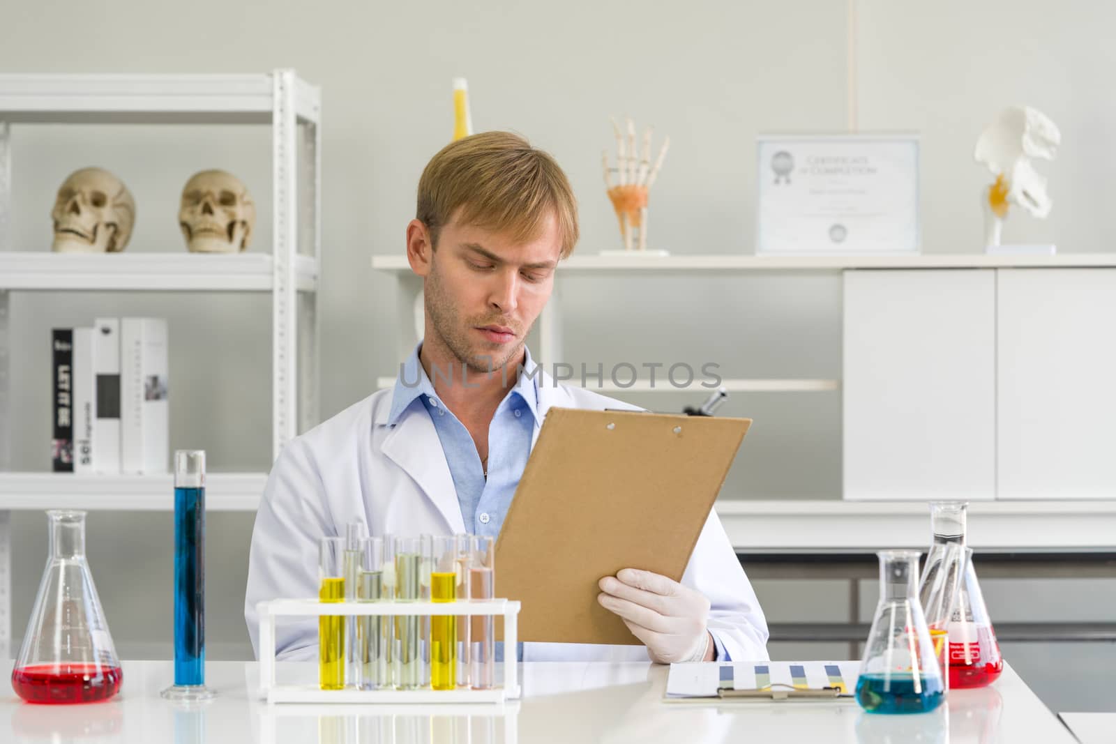 A young Brazilian scientist notes the results of his chemical compound research on the clipboard. Working atmosphere in chemical laboratory. Test tubes filled with chemicals on the table.