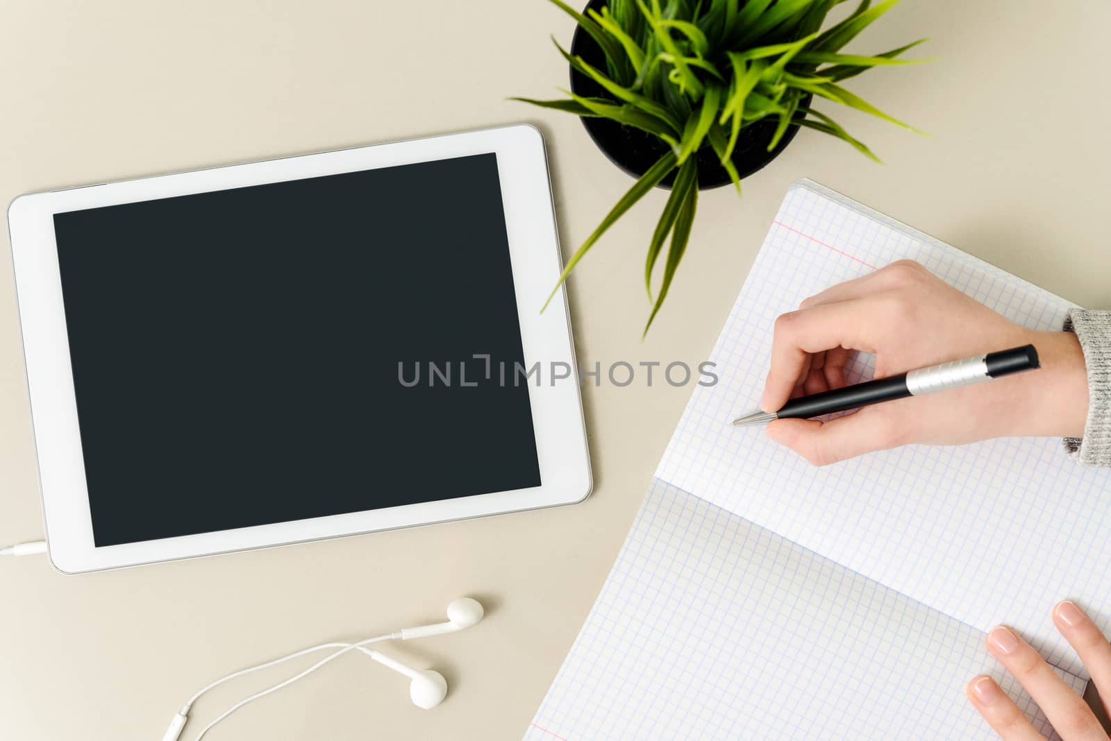 Concept image of student workplace - girl writing a homework in the notebook on the desk next to a digital tablet with blank screen, earphone and flower (high angel view).