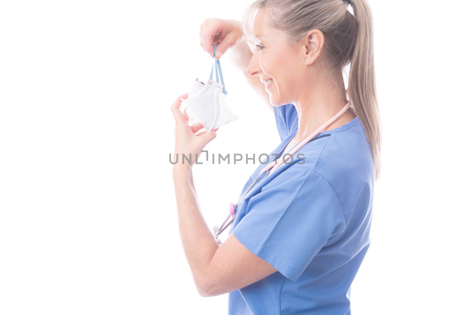 Doctor, nurse or other healthcare worker putting on a respirator N95 face mask.  She is wearing blue scrubs and a stethoscope hangs around her neck. COVID-19 coronavirus or influenza pandemic or other infectios disease