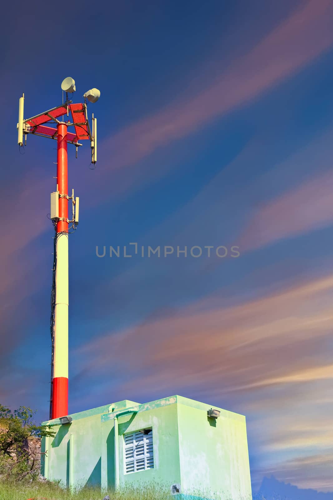 A red and white cell phone tower on a rocky hill by an old shack