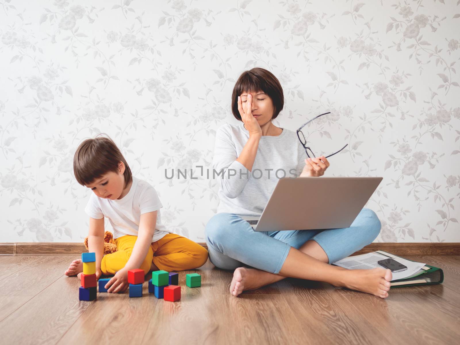 Mom and son sit at home quarantine because of coronavirus COVID19. Tired mother works remotely with laptop, son plays with toy blocks. Self isolation at home.