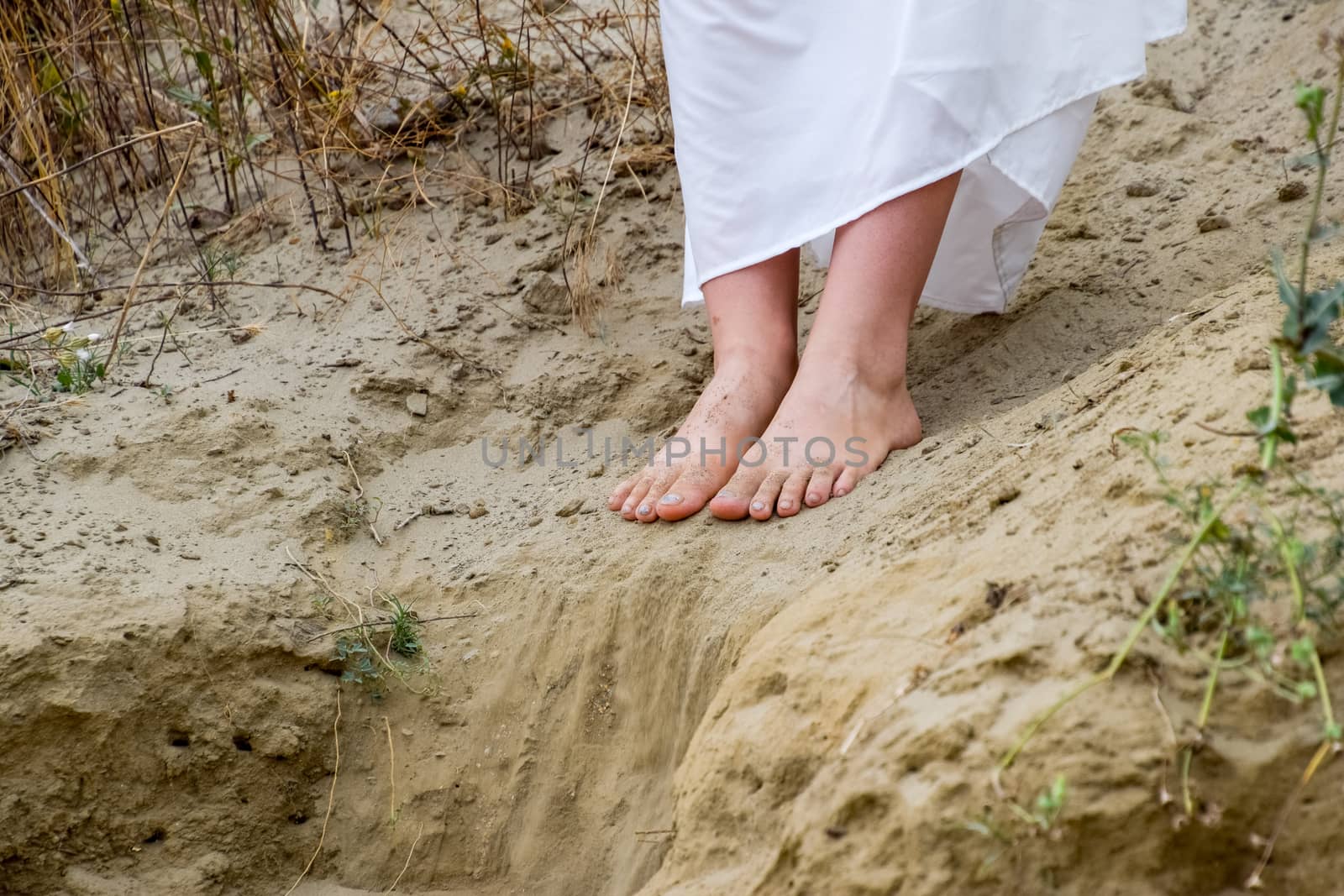 Legs of a girl in a white dress on the edge of a cliff. The sand is falling from the edge of the cliff.