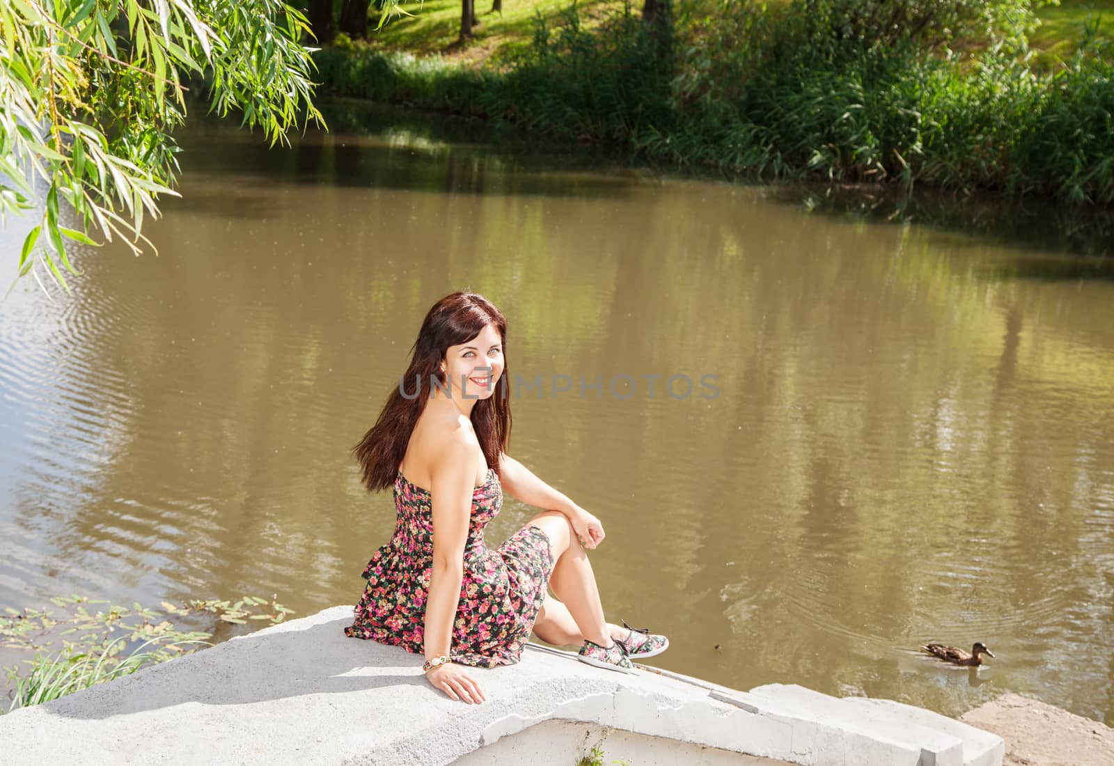 young beautiful smiling brunette sitting near the lake on sunny summer day