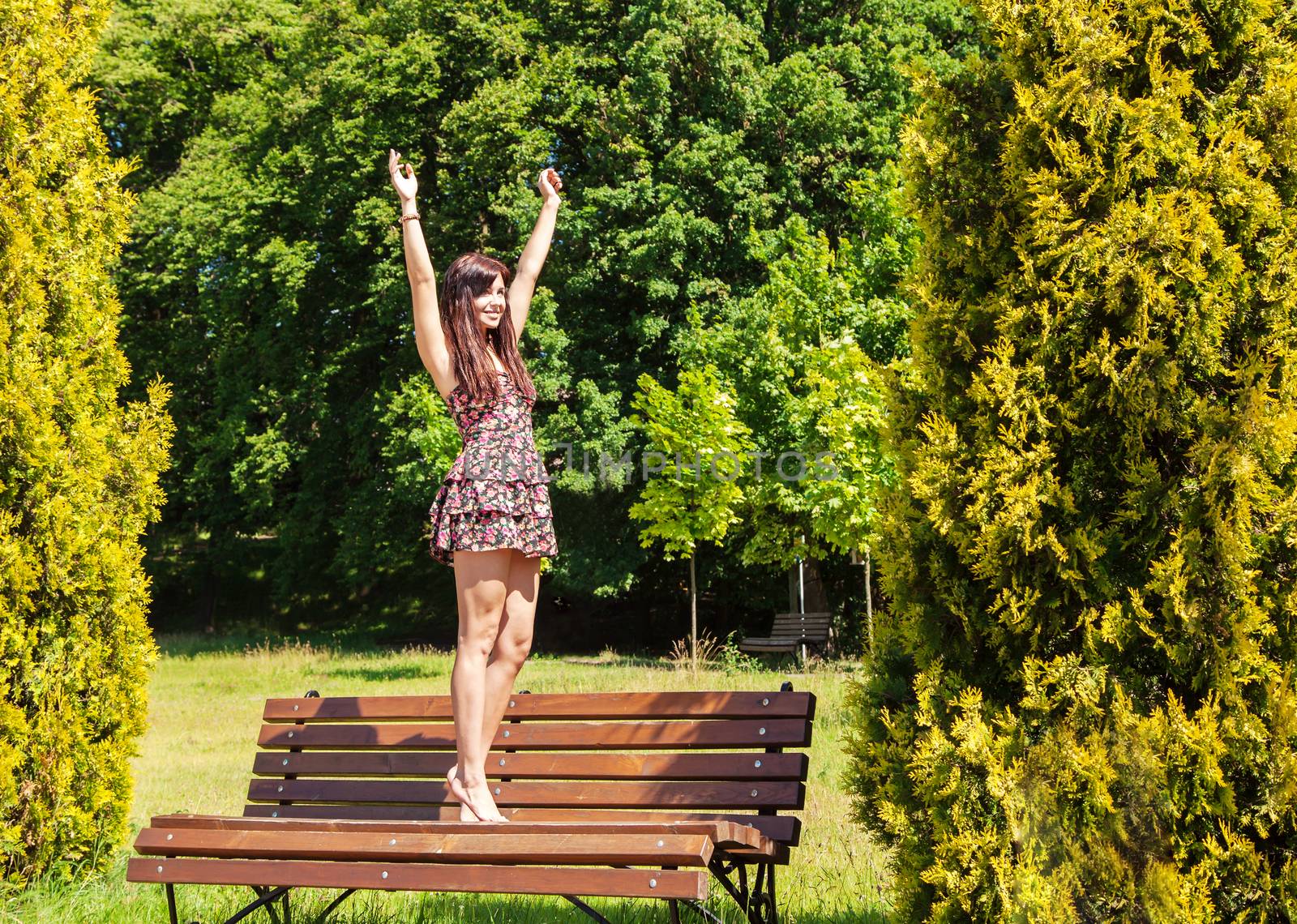 young beautiful smiling brunette standing barefoot on a bench in a city park on sunny summer day