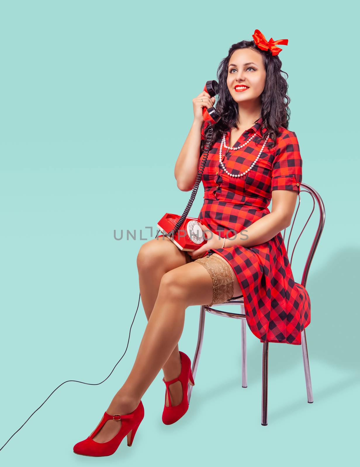 smiling young pinup woman in red dress and nylon stockings sitting on a chair and talking on phone over green background