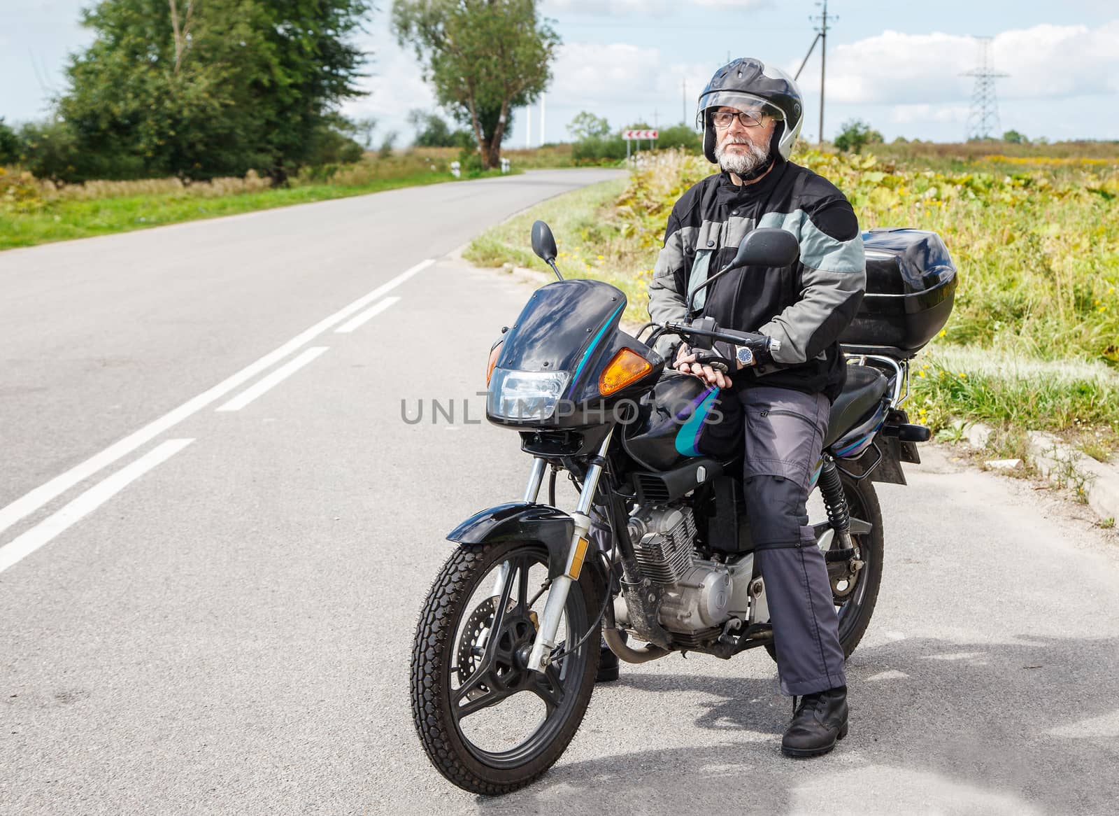 elderly motorcyclist wearing a jacket and glasses with a helmet sitting on his motorcycle on the open road on sunny summer day