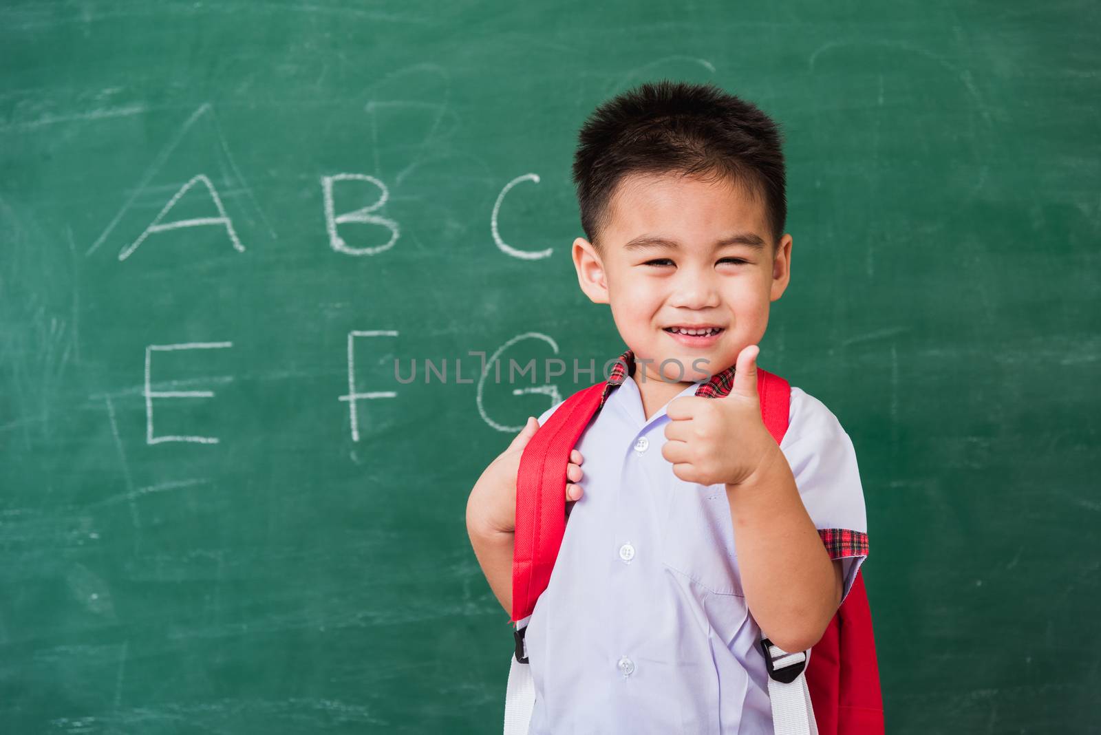 Back to School. Happy Asian funny cute little child boy kindergarten in student uniform with school bag smiling show finger thumb up on green school blackboard, First time to school education concept