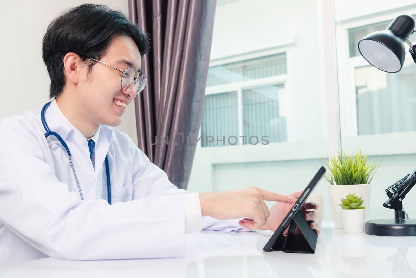 Happy Asian young doctor handsome man smile work from home office wear glasses, using black modern smart digital tablet computer on desk at hospital office, Technology healthcare and medicine concept