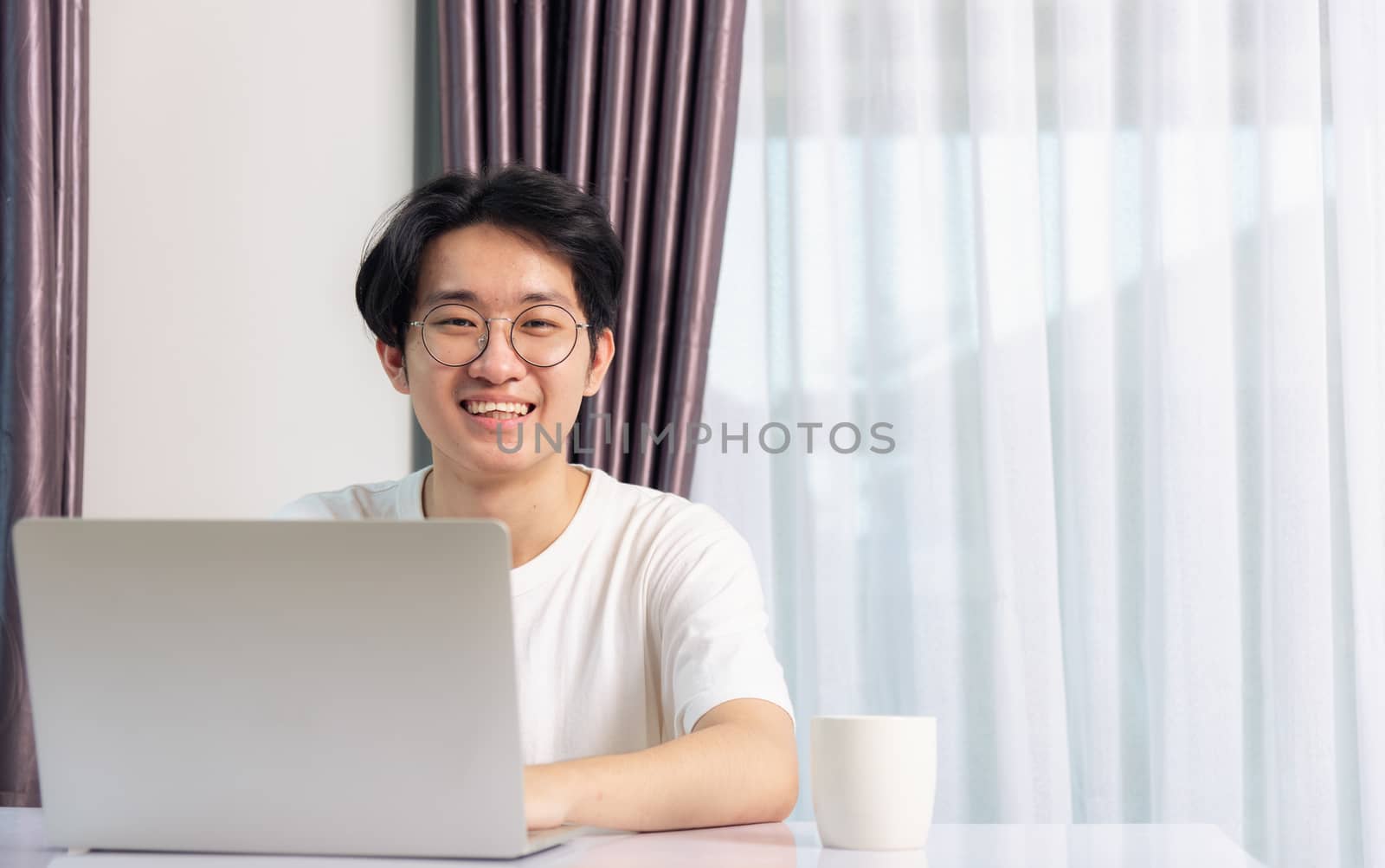 Happy Asian young business handsome man smile work from home office wear glasses and t-shirt comfortable working with laptop computer on desk, Thai person freelance man worker at home