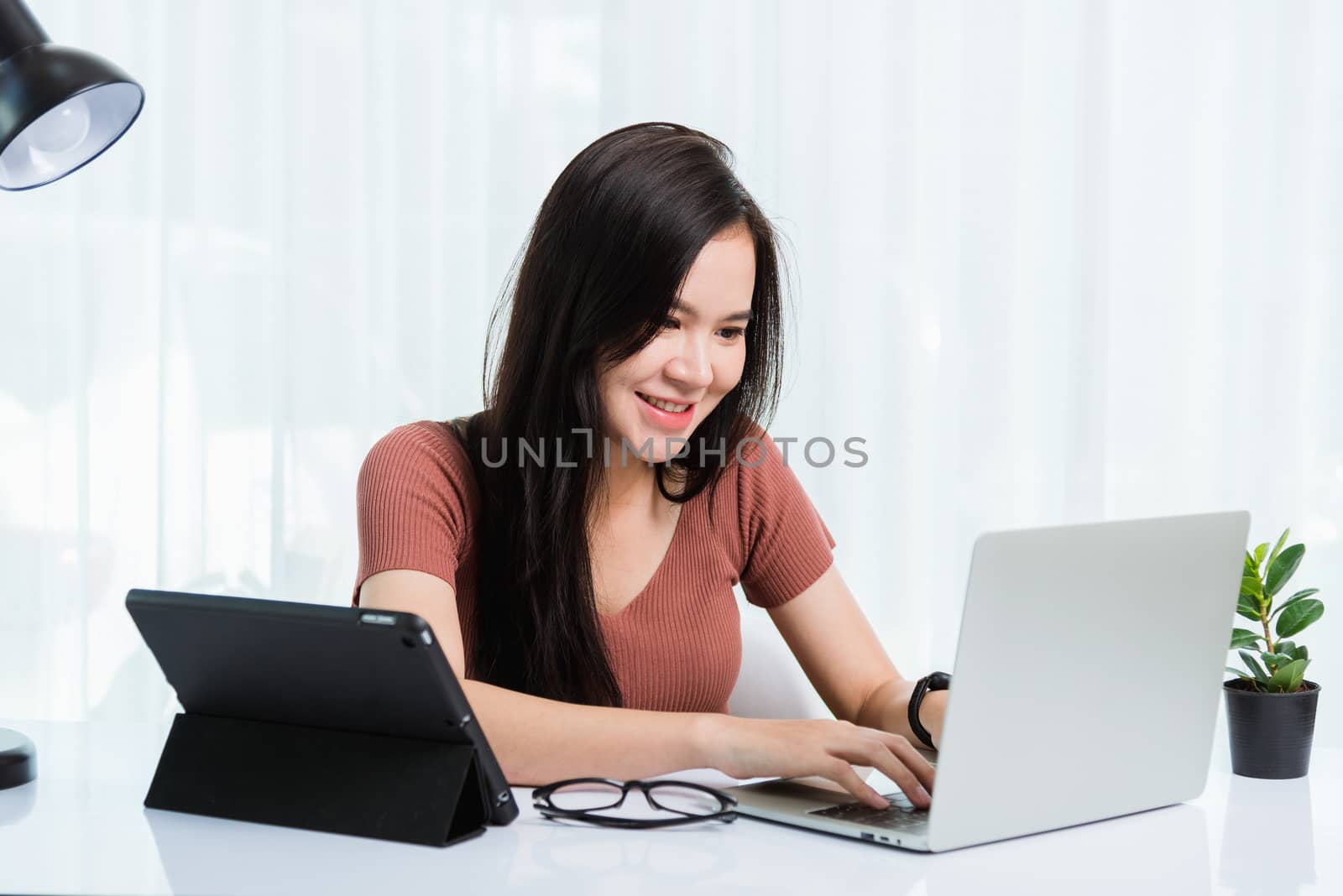 Work from home, Smiling Happy Asian business young beautiful woman sitting on desk workspace use modern smart tablet and laptop computer technology video call conferencing to team at home office