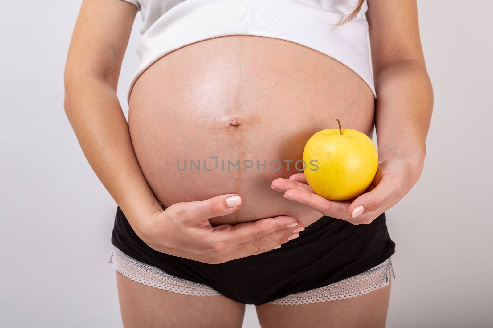 Image of pregnant woman touching her big belly and holding yellow apple in the hand on white background. Close up. Beautiful body of pregnant woman. Motherhood, pregnancy. Woman expecting baby