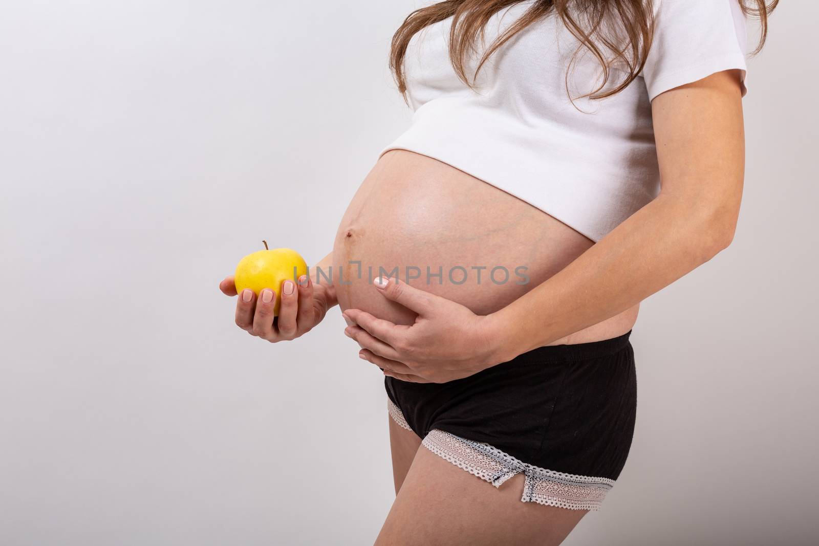 Image of pregnant woman touching her big belly and holding yellow apple in the hand on white background. Close up. Beautiful body of pregnant woman. Motherhood, pregnancy. Woman expecting baby