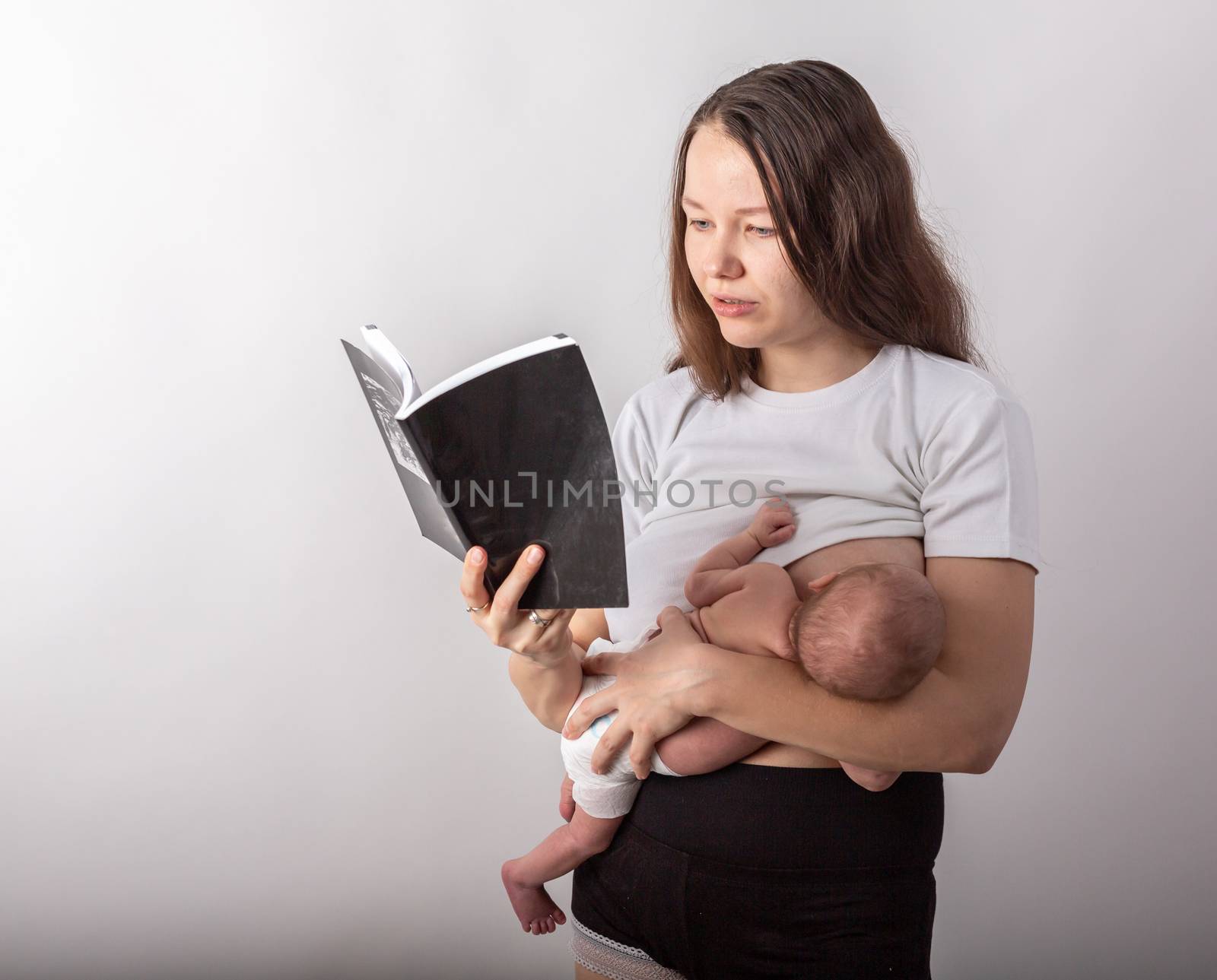 Beautiful young mother breastfeeding a baby while reading a black book by Vassiliy