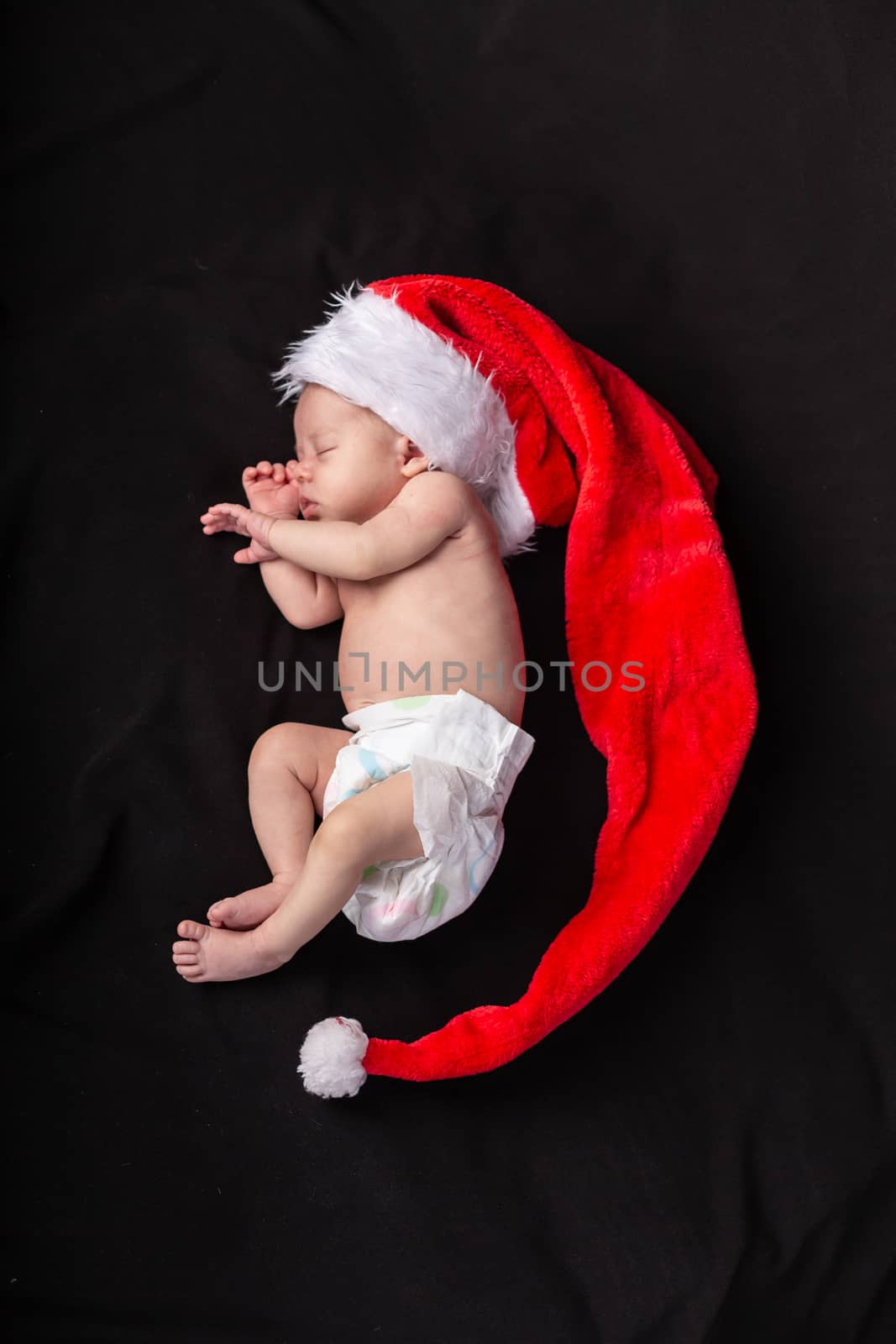 Little baby on a dark background in a long santa claus hat that lies in the shape of a crescent.