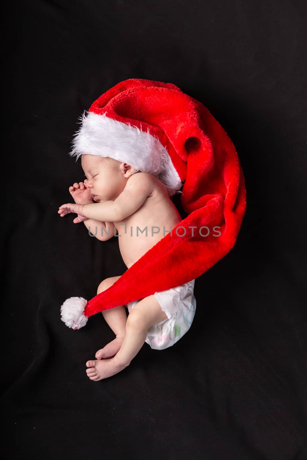 Little baby on a dark background in a long santa claus hat that lies in the shape of a crescent by Vassiliy