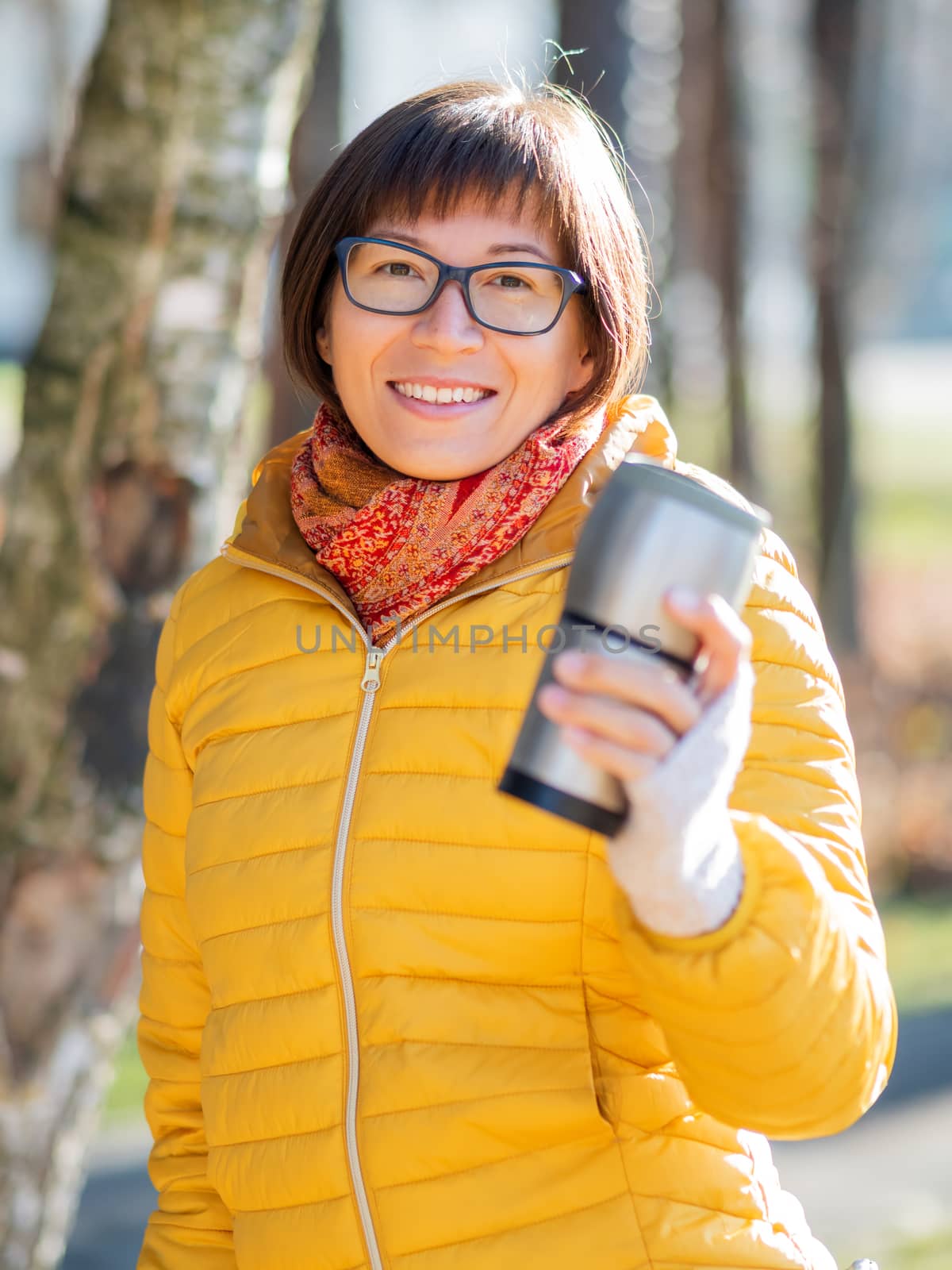 Happy wide smiling women in bright yellow jacket is holding thermos mug. Hot tea or other beverage on cool autumn day.