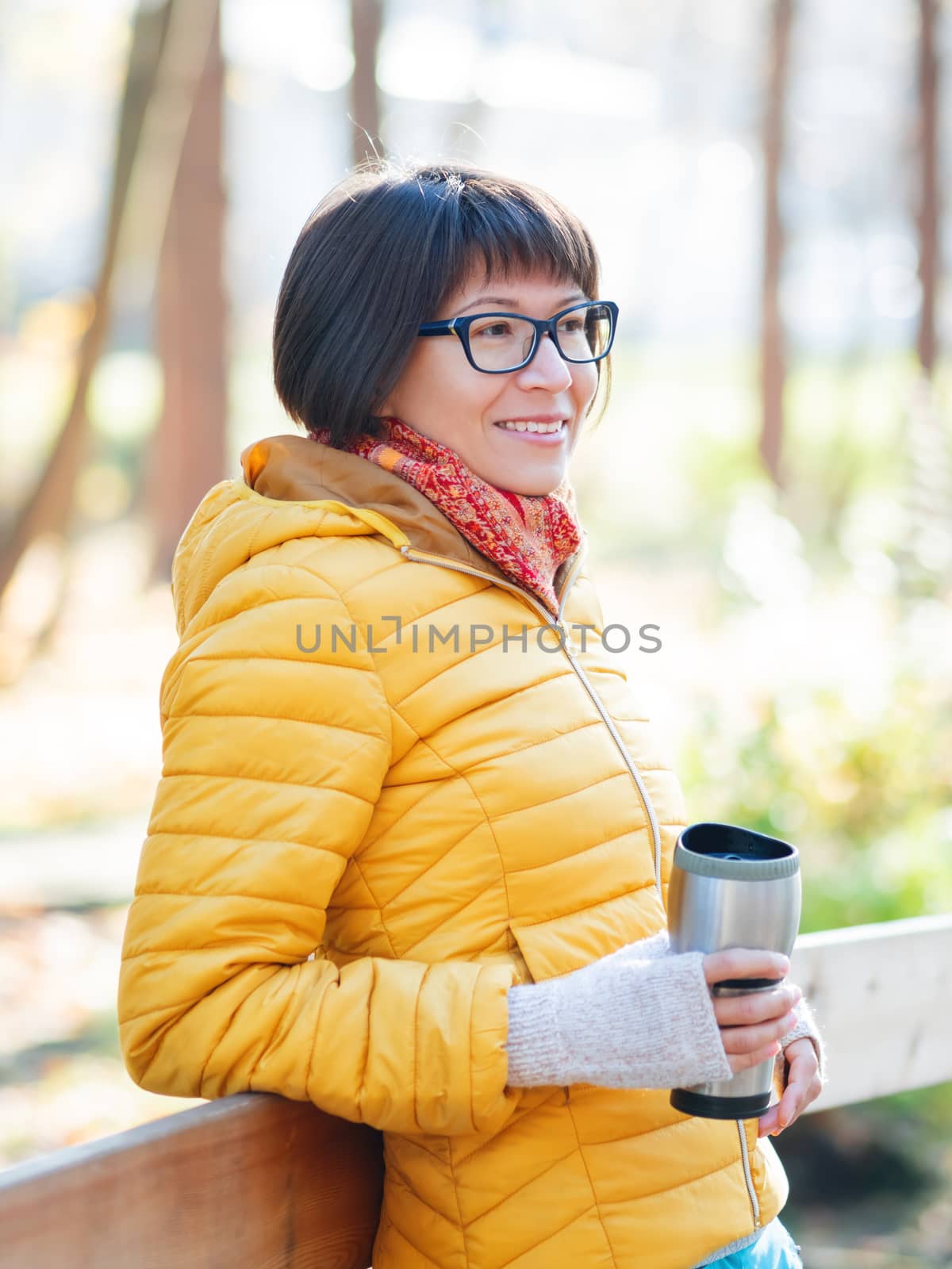 Happy wide smiling women in bright yellow jacket is holding thermos mug. Hot tea or other beverage on cool autumn day.