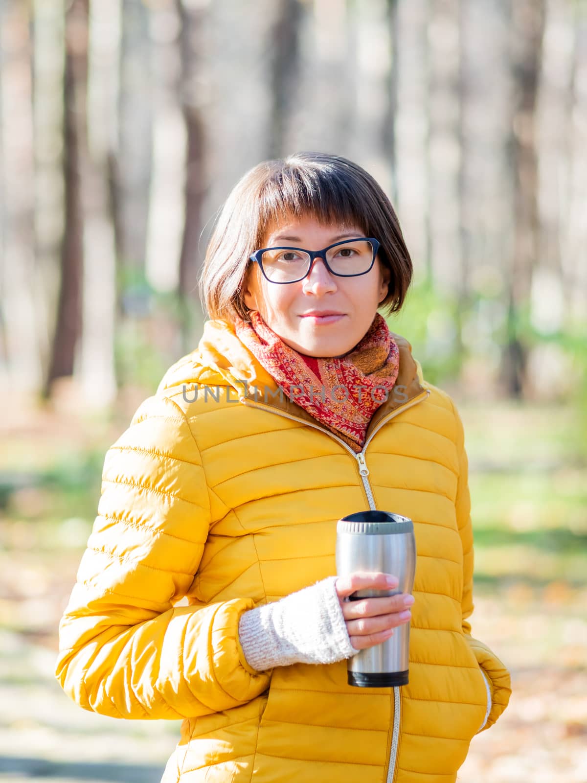 Happy wide smiling women in bright yellow jacket is holding thermos mug. Hot tea or other beverage on cool autumn day.