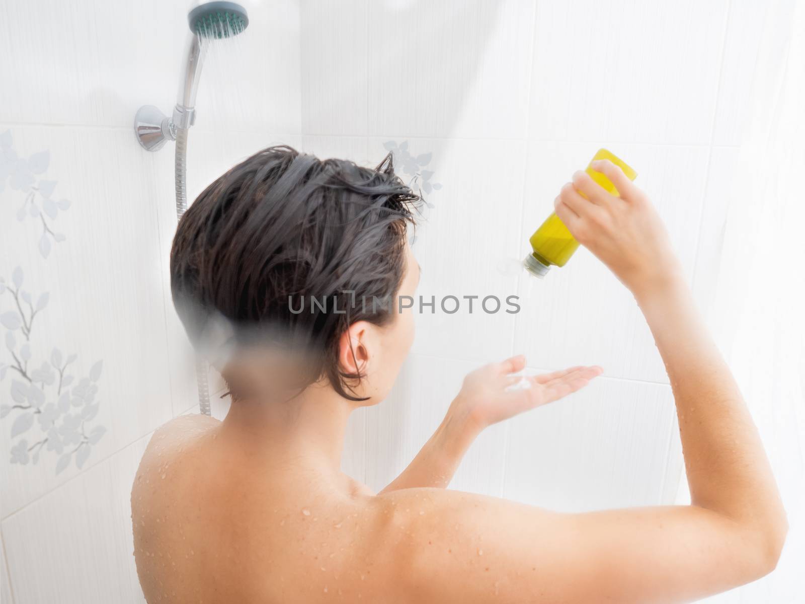 Naked woman with short hair takes a shower. Woman squeezes shampoo out of a green bottle. White bathroom.