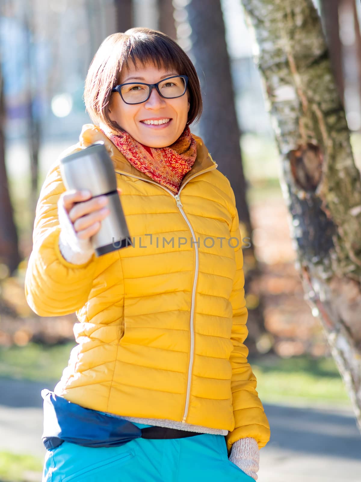 Happy wide smiling women in bright yellow jacket is holding thermos mug. Hot tea or other beverage on cool autumn day. by aksenovko