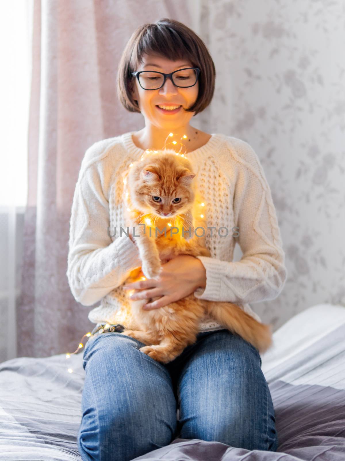 Cute ginger cat tangled in light bulb garland. Woman in knitted sweater trying to make fluffy pet free. Christmas decorations. Cozy home before New Year.