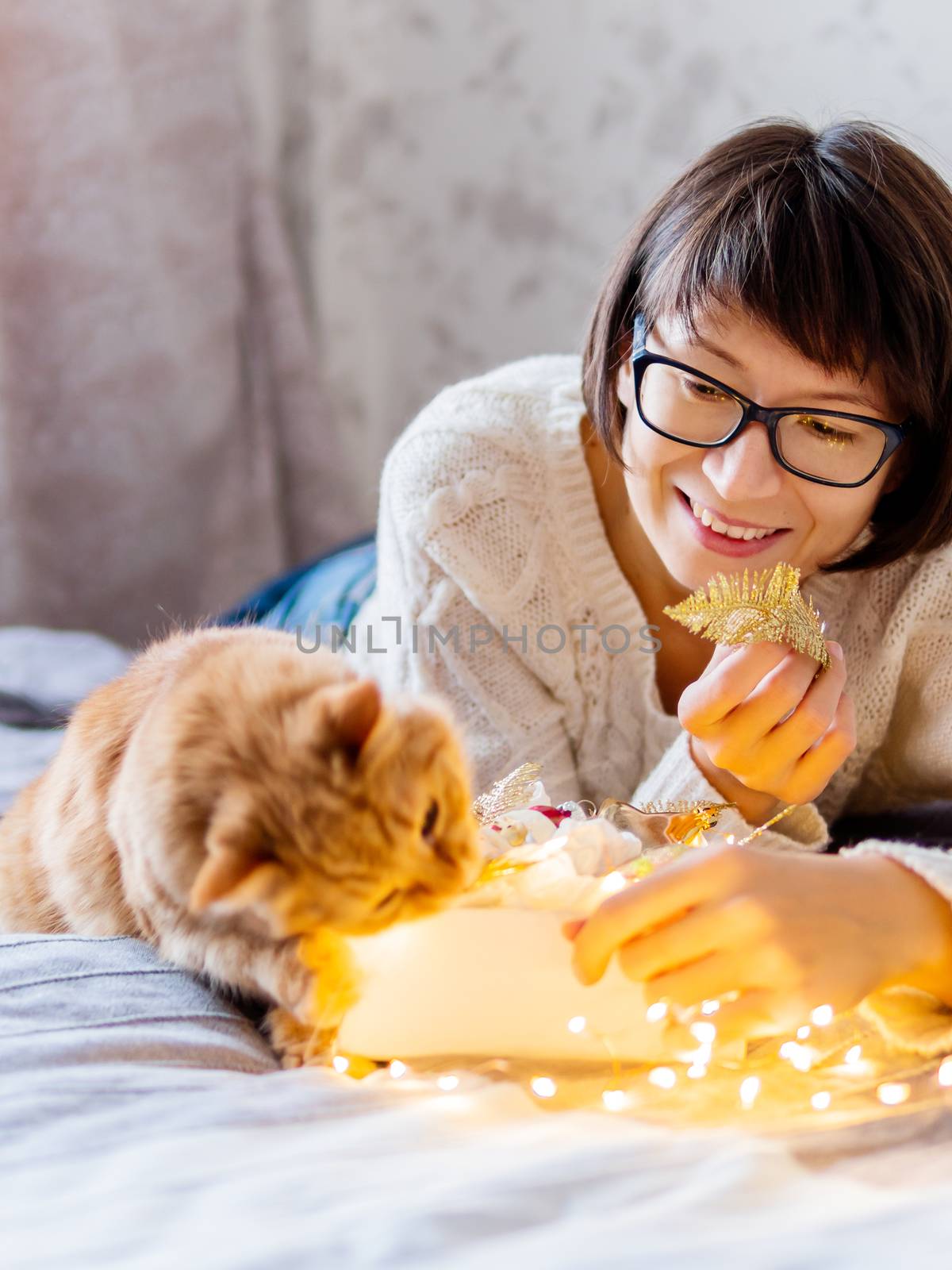 Cute ginger cat with bites light bulbs. Woman playing with her fluffy pet and box with Christmas decorations. Cozy home before New Year.