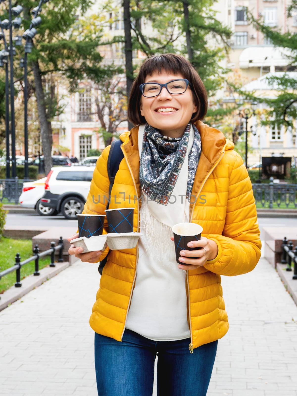 Happy wide smiling women in bright yellow jacketis holding paper cup with hot coffee. Hot beverage on cool autumn day. by aksenovko