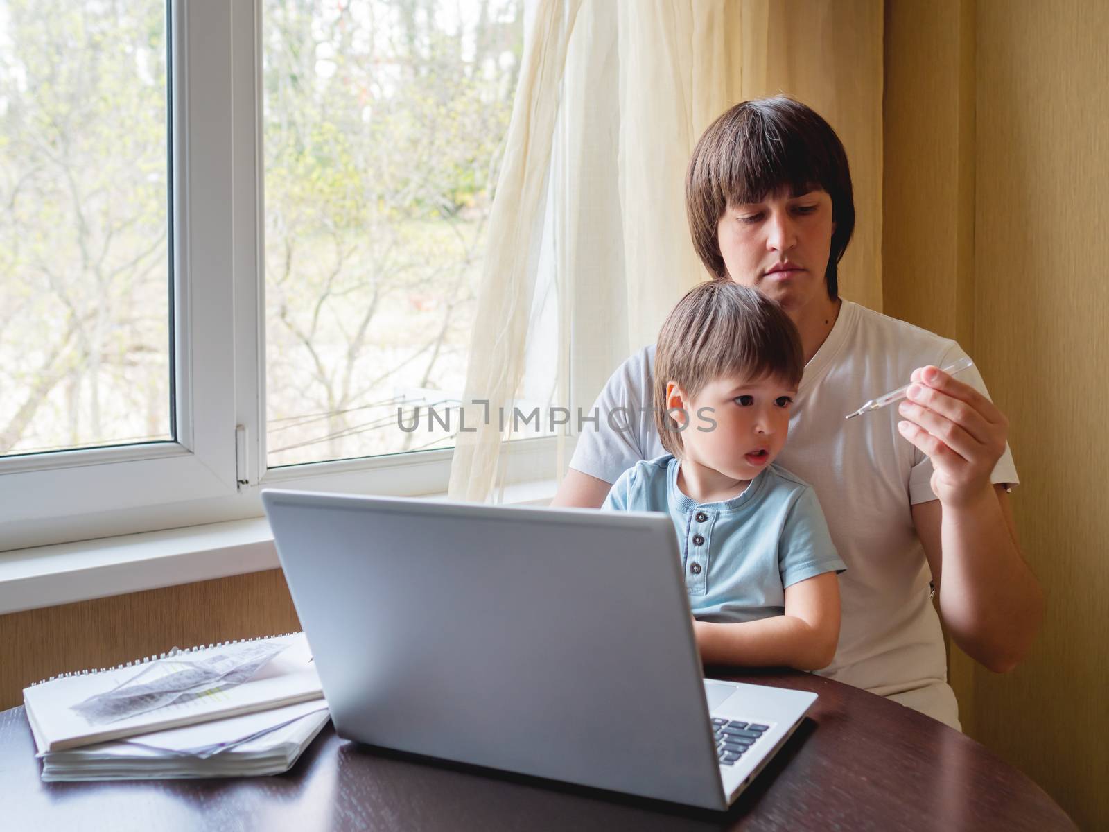 Self-isolation because of coronavirus outbreak. Man is remote working with his little son. Dad looks at the thermometer. Laptop on table. by aksenovko