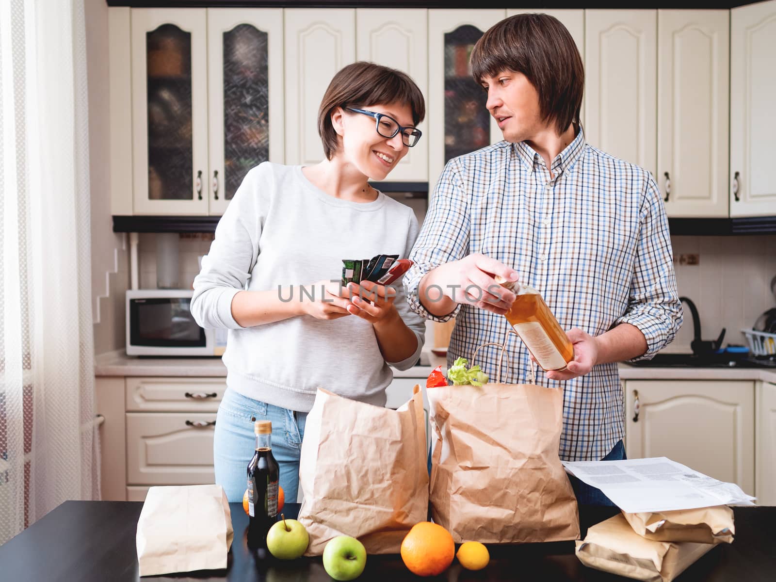 Young couple is sorting out purchases in the kitchen. Products in bags made of craft paper. Food delivery in conditions of quarantine because of coronavirus COVID19.