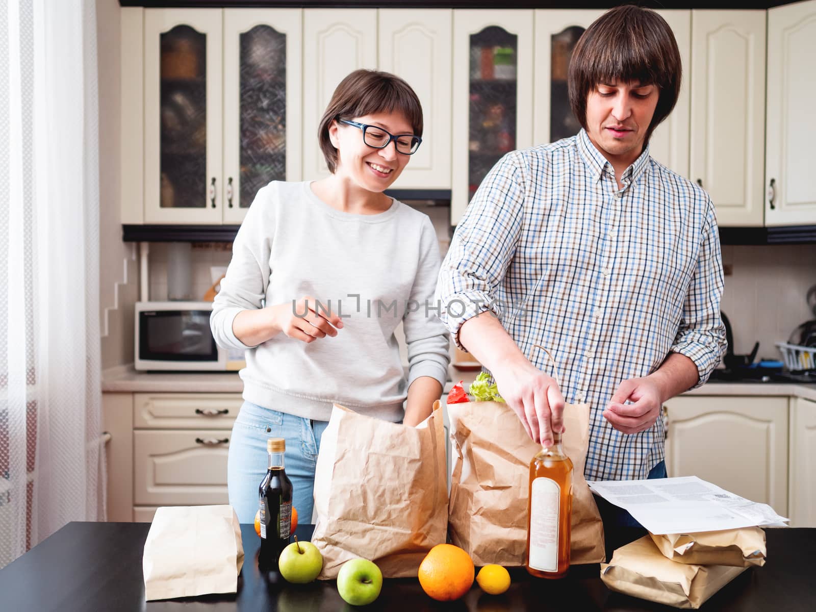 Young couple is sorting out purchases in the kitchen. Products in bags made of craft paper. Food delivery in conditions of quarantine because of coronavirus COVID19. by aksenovko