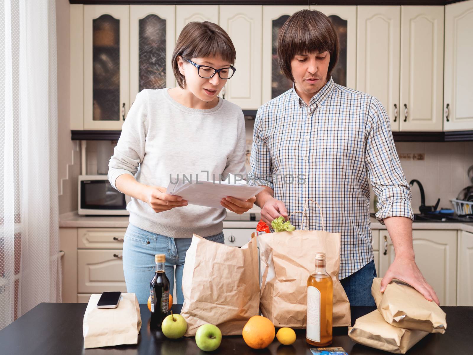 Young couple is sorting out purchases in the kitchen. Products in bags made of craft paper. Food delivery in conditions of quarantine because of coronavirus COVID19.