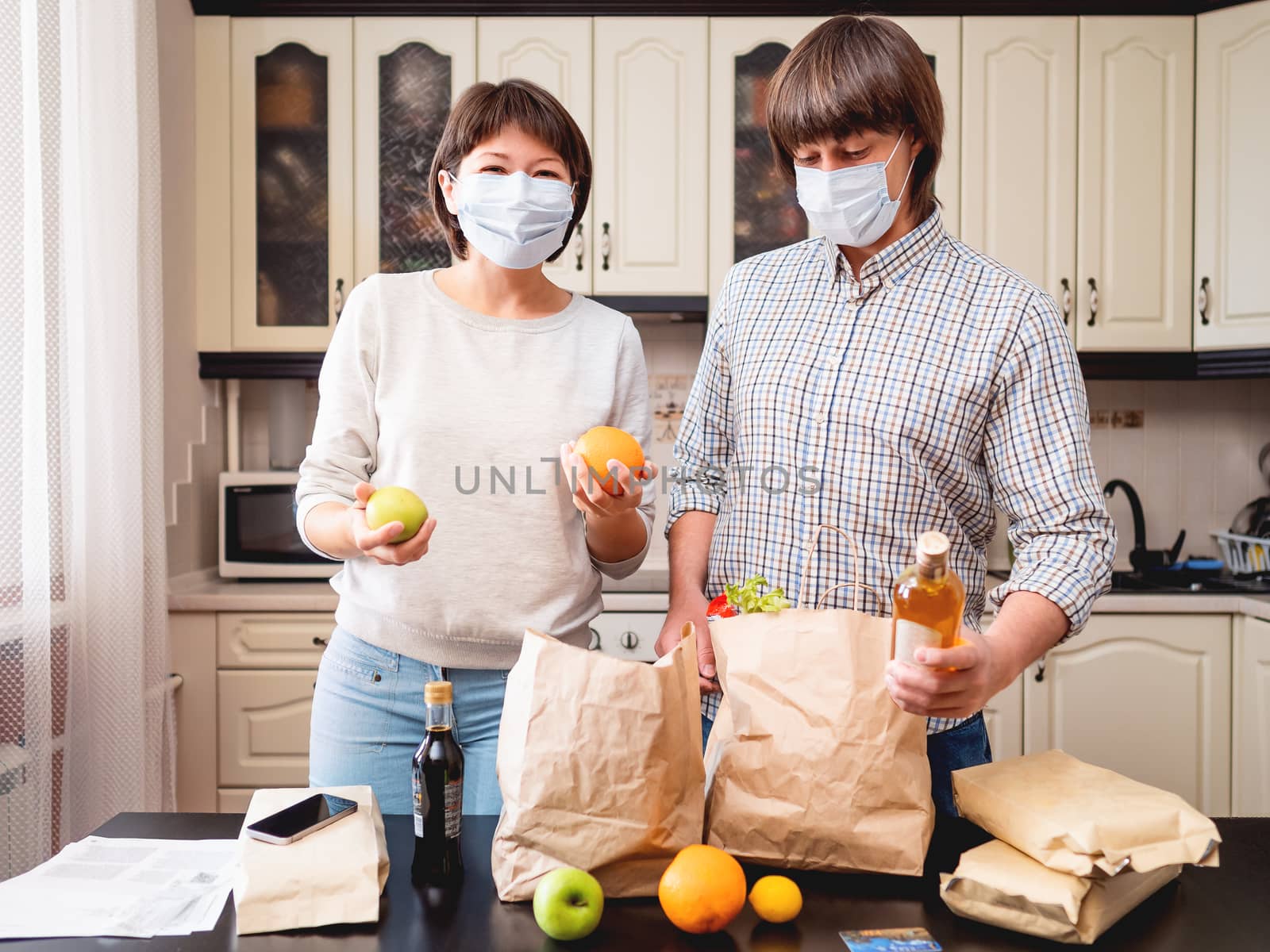 Young couple in protective masks is sorting out purchases in the kitchen. Products in bags made of craft paper. Food delivery in conditions of quarantine because of coronavirus COVID19. by aksenovko