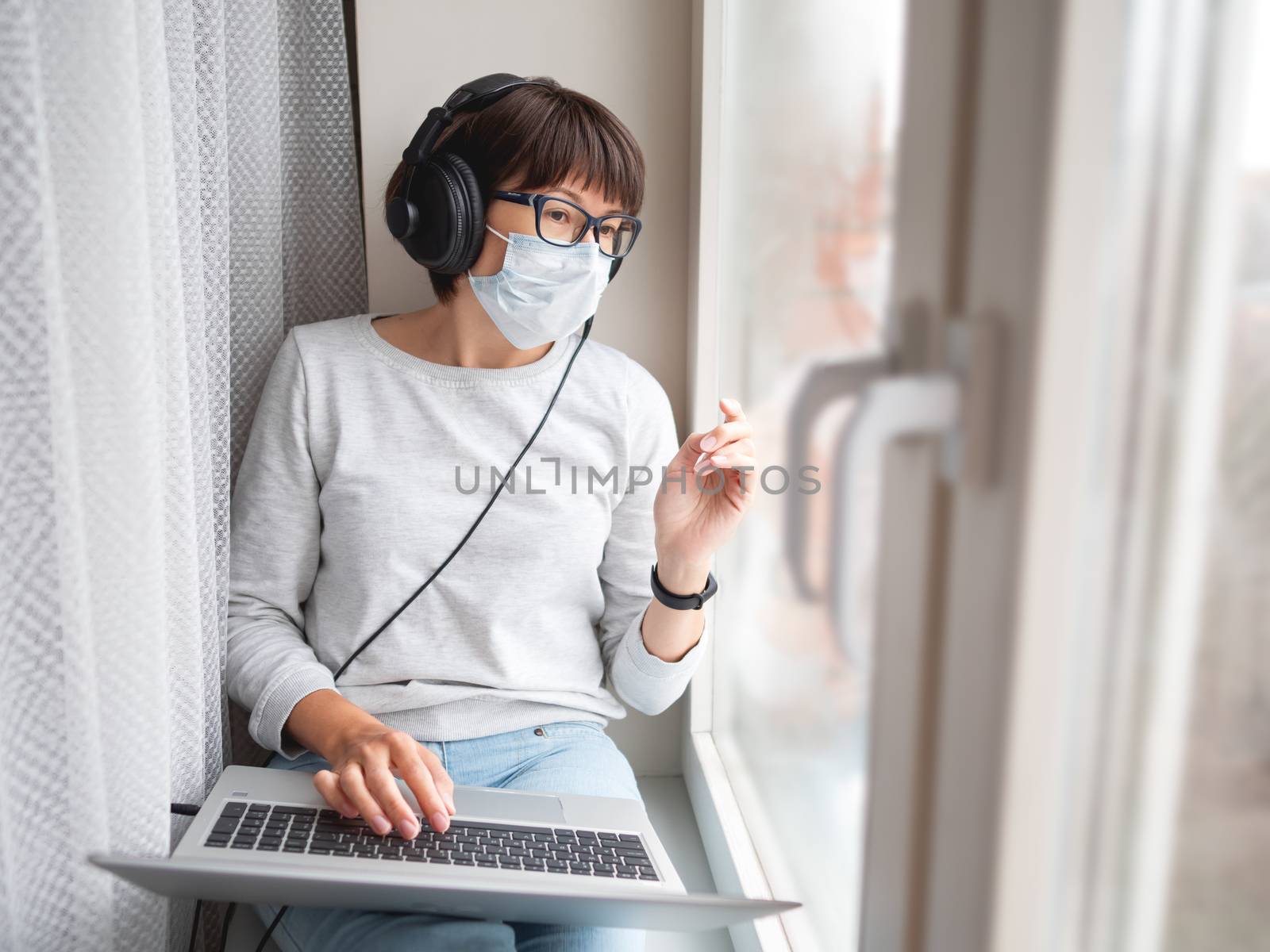 Woman in medical mask remote works from home. She sits on window sill with laptop on knees and headphones. Lockdown quarantine because of coronavirus COVID19. Self isolation at home.