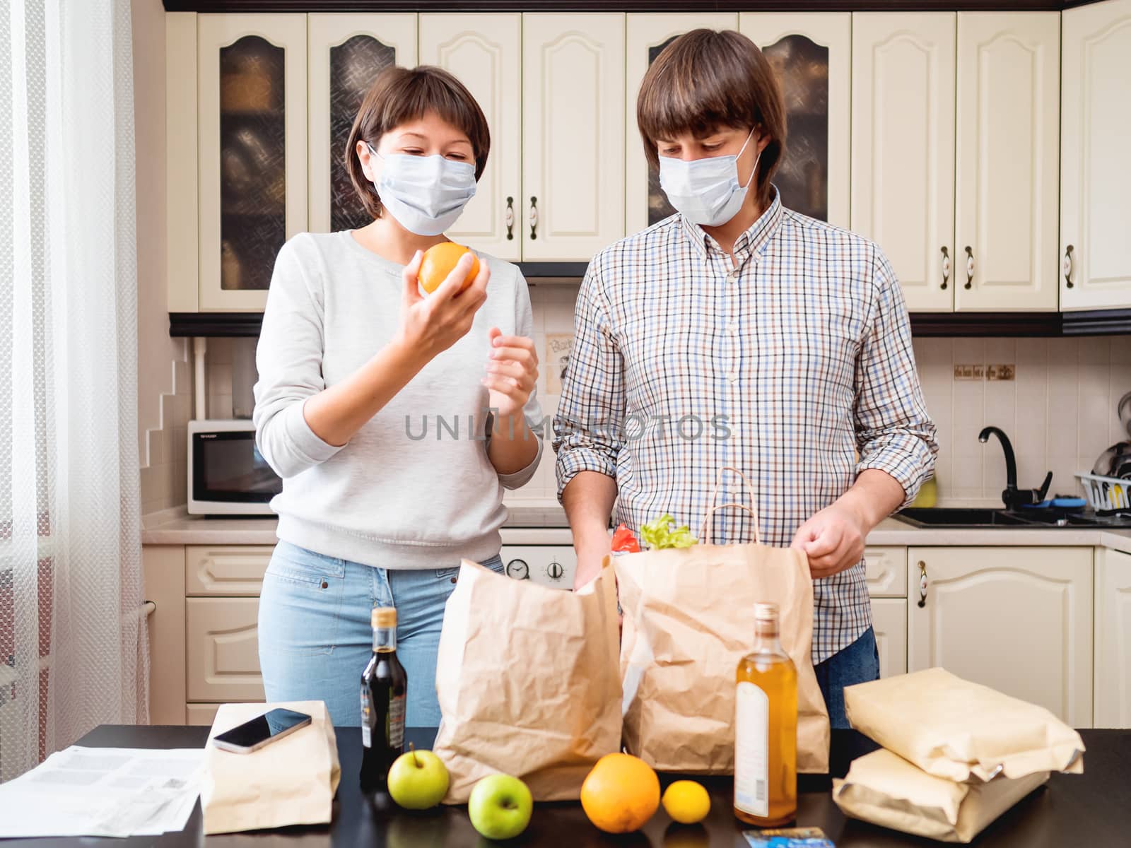 Young couple in protective masks is sorting out purchases in the kitchen. Products in bags made of craft paper. Food delivery in conditions of quarantine because of coronavirus COVID19. by aksenovko