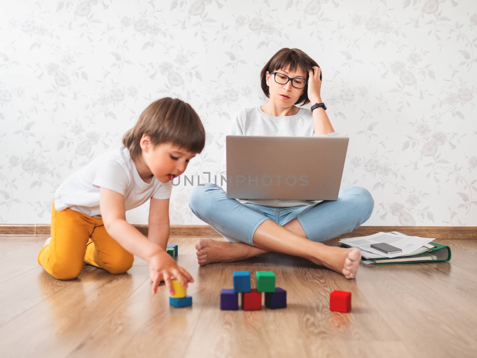 Mom and son sitting at home quarantine because of coronavirus COVID19. Mother remote works with laptop, son plays with toy blocks. Self isolation at home.