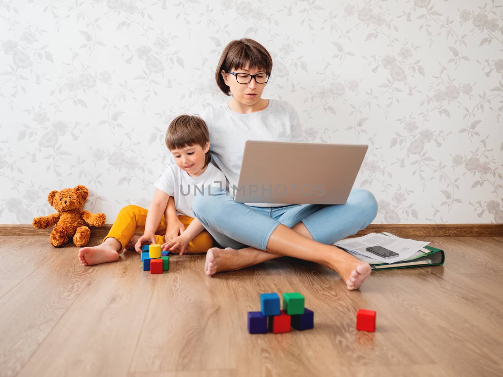 Mom and son sitting at home quarantine because of coronavirus COVID19. Mother remote works with laptop, son plays with toy blocks. Self isolation at home. by aksenovko