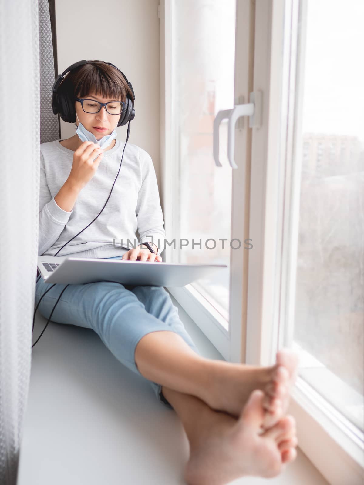 Woman in medical mask remote works from home. She sits on window sill with laptop on knees and headphones. Lockdown quarantine because of coronavirus COVID19. Self isolation at home.