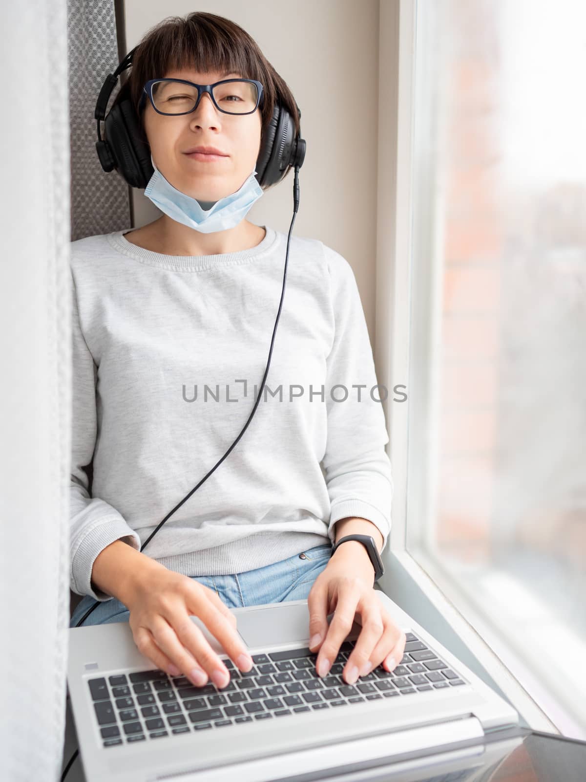 Woman in medical mask remote works from home. She sits on window sill with laptop on knees and headphones. Lockdown quarantine because of coronavirus COVID19. Self isolation at home. by aksenovko