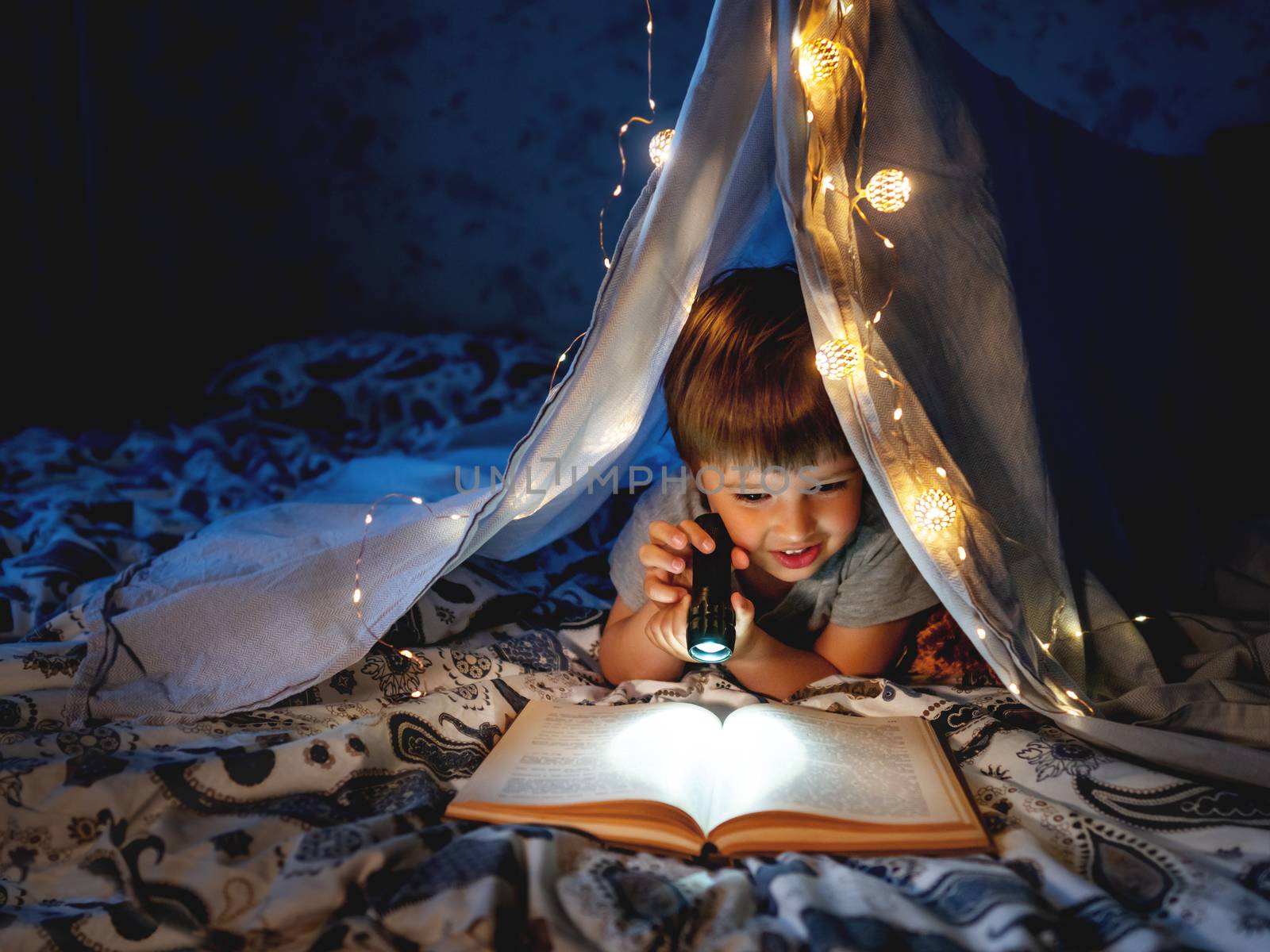 Little boy reads with pocket light. Toddler plays in tent made of linen sheet on bed. Cozy evening with favorite book.