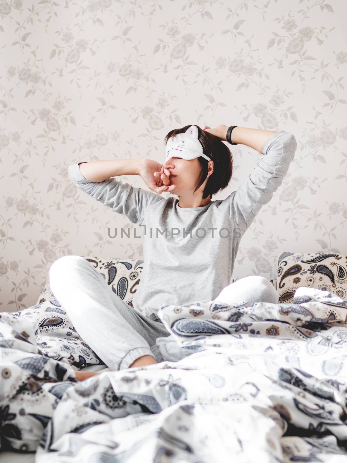 Young woman in grey pajama and sleeping mask in shape of cute sleeping cat face. She is just woke up and sit in bed. Early morning in cozy home. by aksenovko