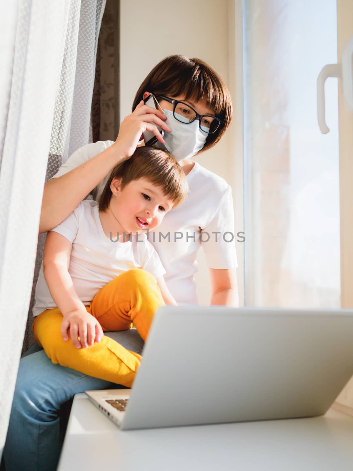 Woman in medical mask remote works from home. She sits on window sill with laptop and cute toddler boy on her knees. Lockdown quarantine because of coronavirus COVID19. Self isolation at home.