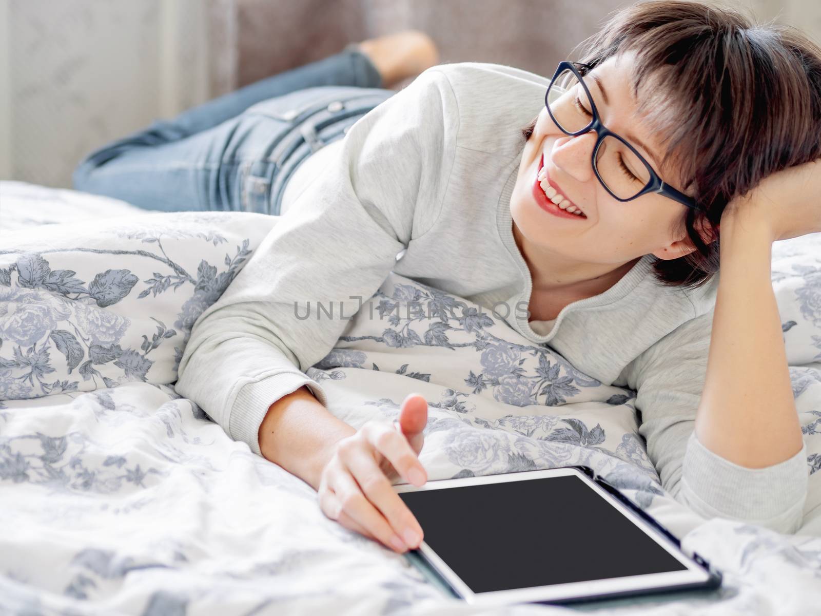 Woman in glasses are lying in bed with tablet. She is touching screen. Morning bedtime.