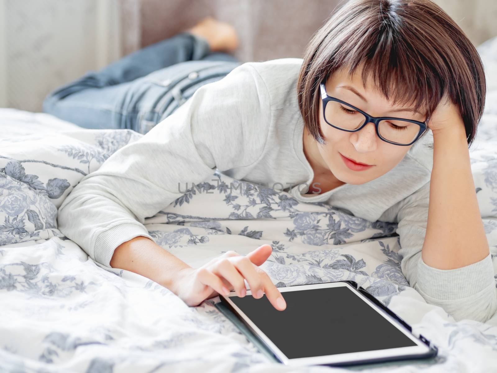 Woman in glasses are lying in bed with tablet. She is touching screen. Morning bedtime.