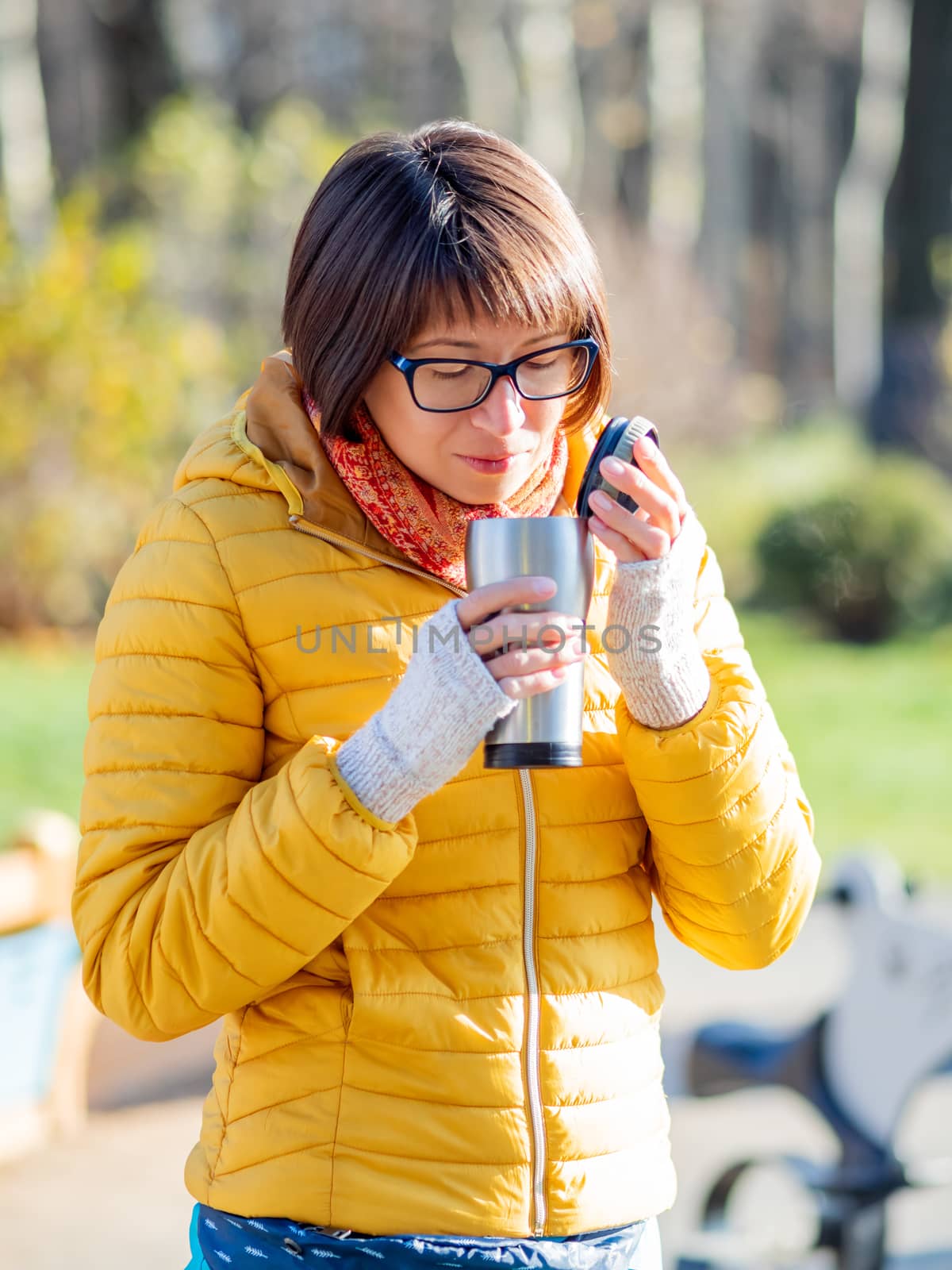 Happy wide smiling women in bright yellow jacket is holding thermos mug. Hot tea or other beverage on cool autumn day.