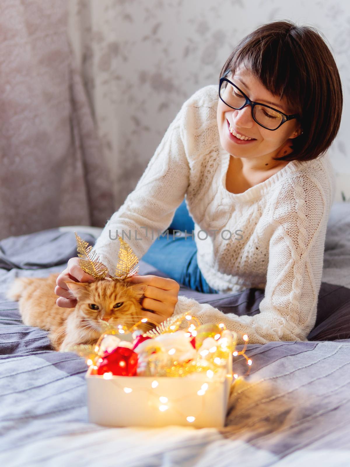 Cute ginger cat with bright golden ears made of decorative feathers. Woman playing with her fluffy pet and box with Christmas decorations. Cozy home before New Year.