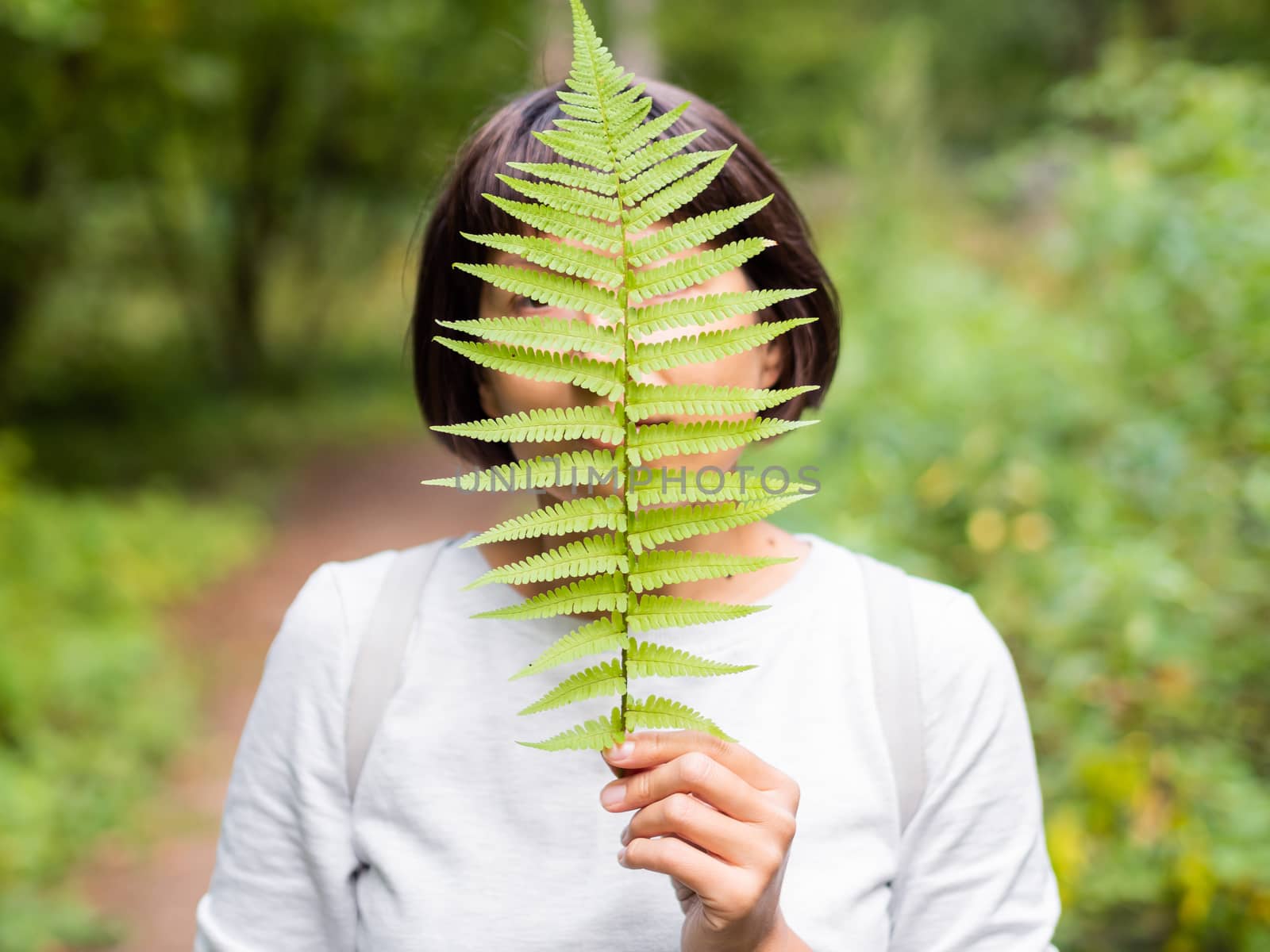 Young woman is hiding her eyes with fern leaf. Symbol of life, tranquility and unity with nature. Summer in forest.