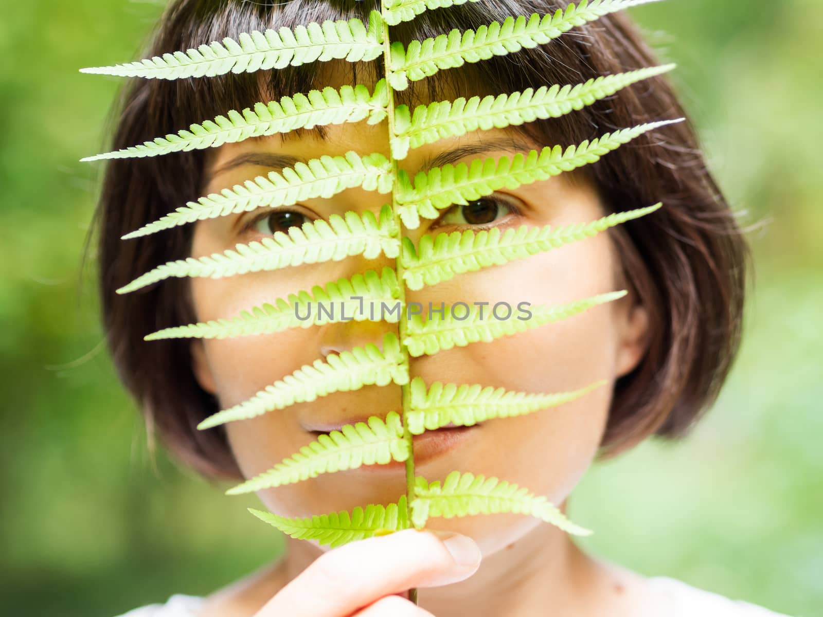 Young woman is hiding her eyes with fern leaf. Symbol of life, tranquility and unity with nature. Summer in forest.