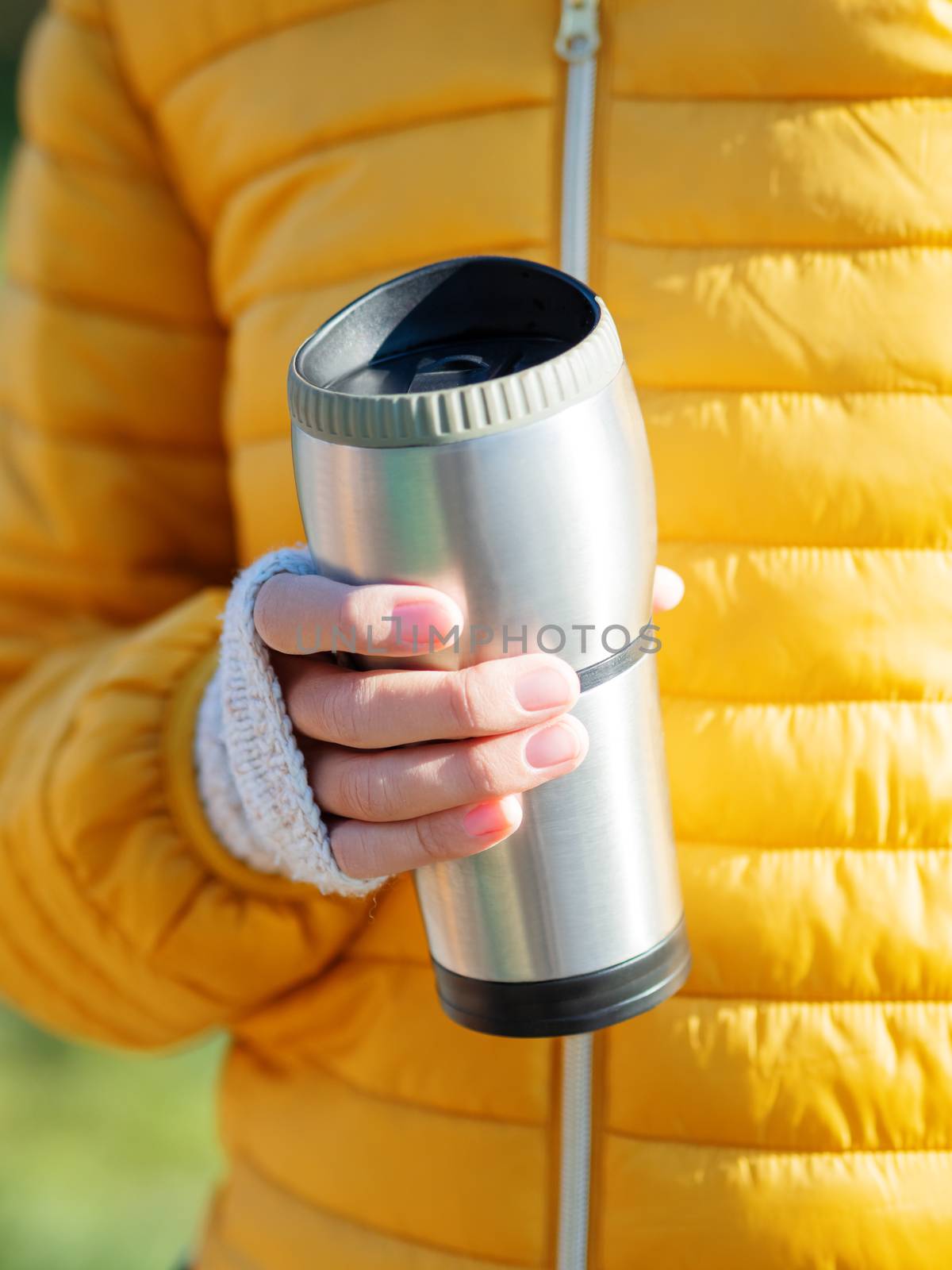 Women in bright yellow jacket is holding thermos mug. Hot tea or other beverage on cool autumn day.