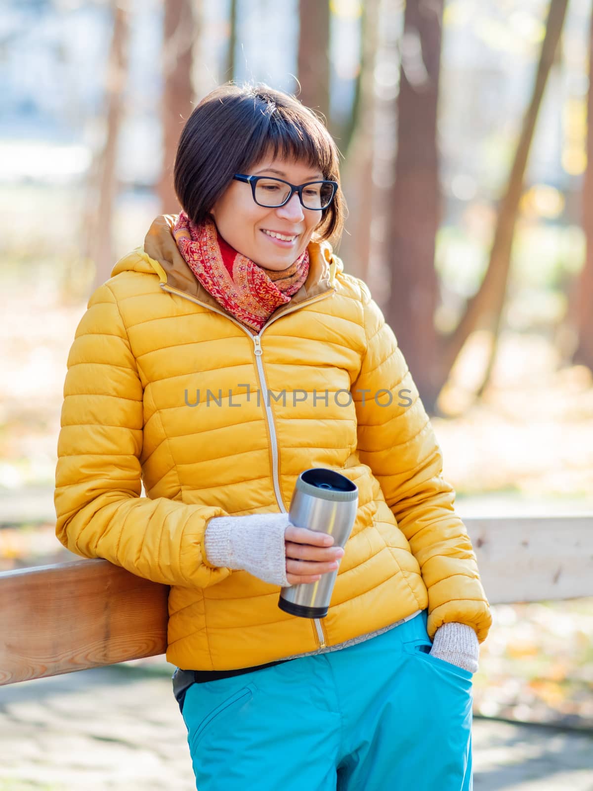 Happy wide smiling women in bright yellow jacket is holding thermos mug. Hot tea or other beverage on cool autumn day.